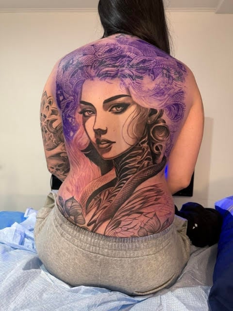 Back view of a woman with a large purple and pink portrait tattoo on her back and decorative arm tattoos
