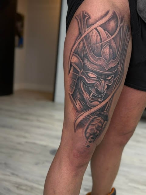 Close-up of a tattoo on a mans leg featuring a detailed demon or oni mask with horns and intricate designs in black ink