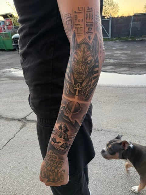 Person wearing black pants showing full sleeve Egyptian-themed tattoo covering arm and leg, standing outdoors with dog nearby