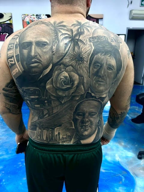 Man with large back tattoo featuring multiple portrait faces, palm trees, roses, and architectural elements in black and gray ink