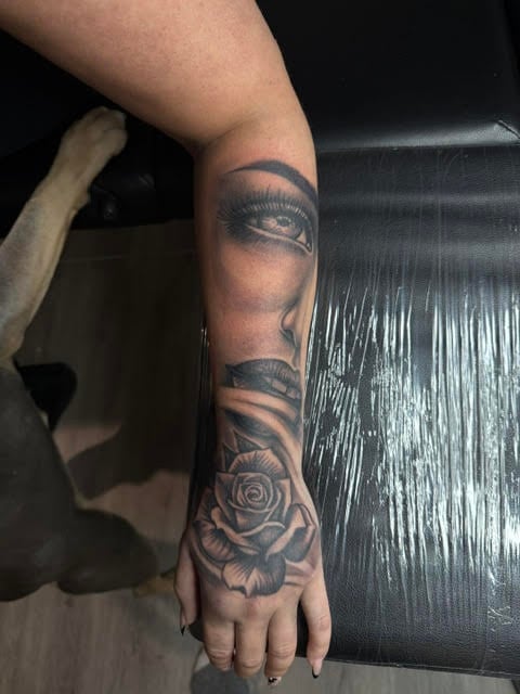 Arm with black and gray tattoo of female face with closed eyes and rose flower on hand