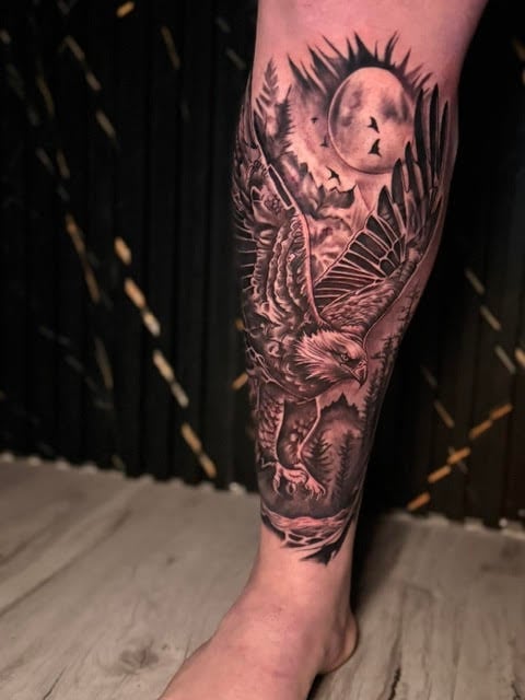 Detailed black and gray leg tattoo featuring a moon, wings, butterfly, flowers, and nature elements on a calf